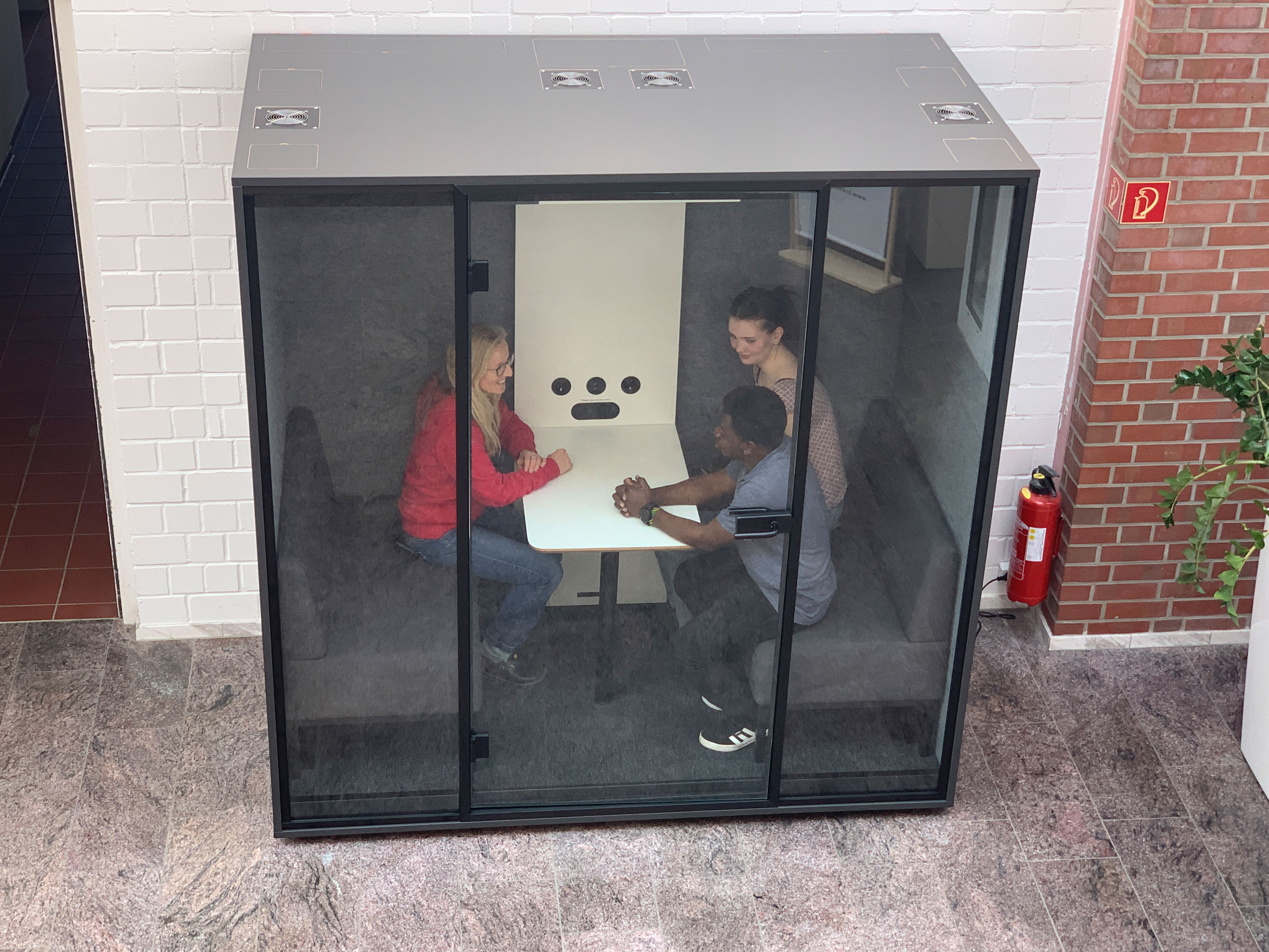 Drei Personen sitzen in einer kleinen, gläsernen Besprechungskabine mit dunklen Polsterbänken und einem weißen Tisch. Zwei Personen sitzen sich gegenüber und beugen sich leicht nach vorne im Gespräch, während eine dritte Person seitlich sitzt und aufmerksam teilnimmt. 