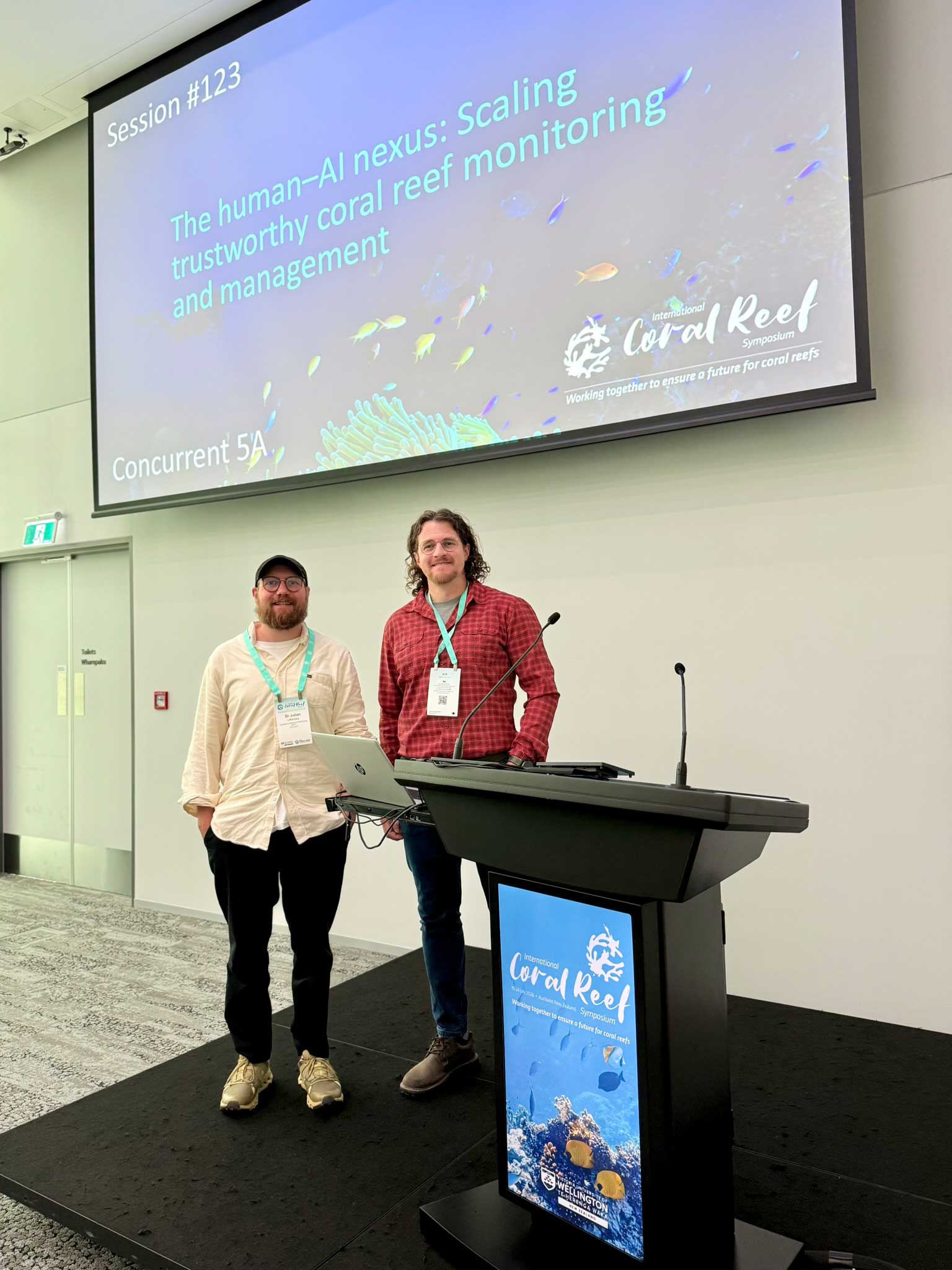 Zwei Männer stehen für ein Foto neben einem Rednerpult. Über ihnen zeigt eine große Leinwand die Sitzung „The human–AI nexus: Scaling trustworthy coral reef monitoring and management“ beim International Coral Reef Symposium.