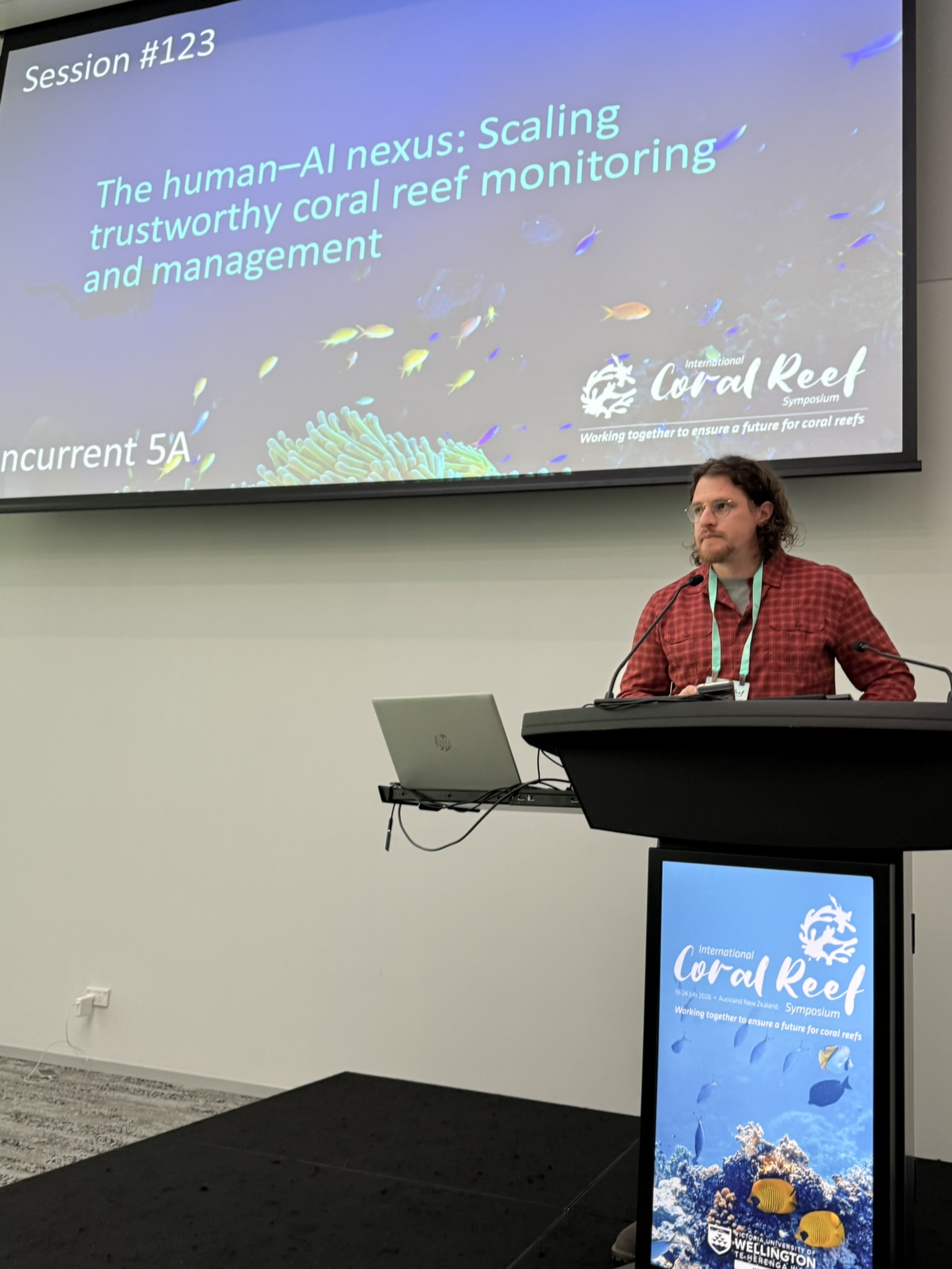 Ein Mann spricht an einem Rednerpult während einer Konferenzsitzung. Auf der großen Leinwand hinter ihm steht „The human–AI nexus: Scaling trustworthy coral reef monitoring and management“.