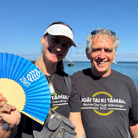 Das Foto zeigt eine Frau und einen Mann in grauen T-Shirts im Sonneschein vor blauem Himmel. Die Frau hält einen blauen Papierfächer mit der Aufschrift ZMT in die Kamera.