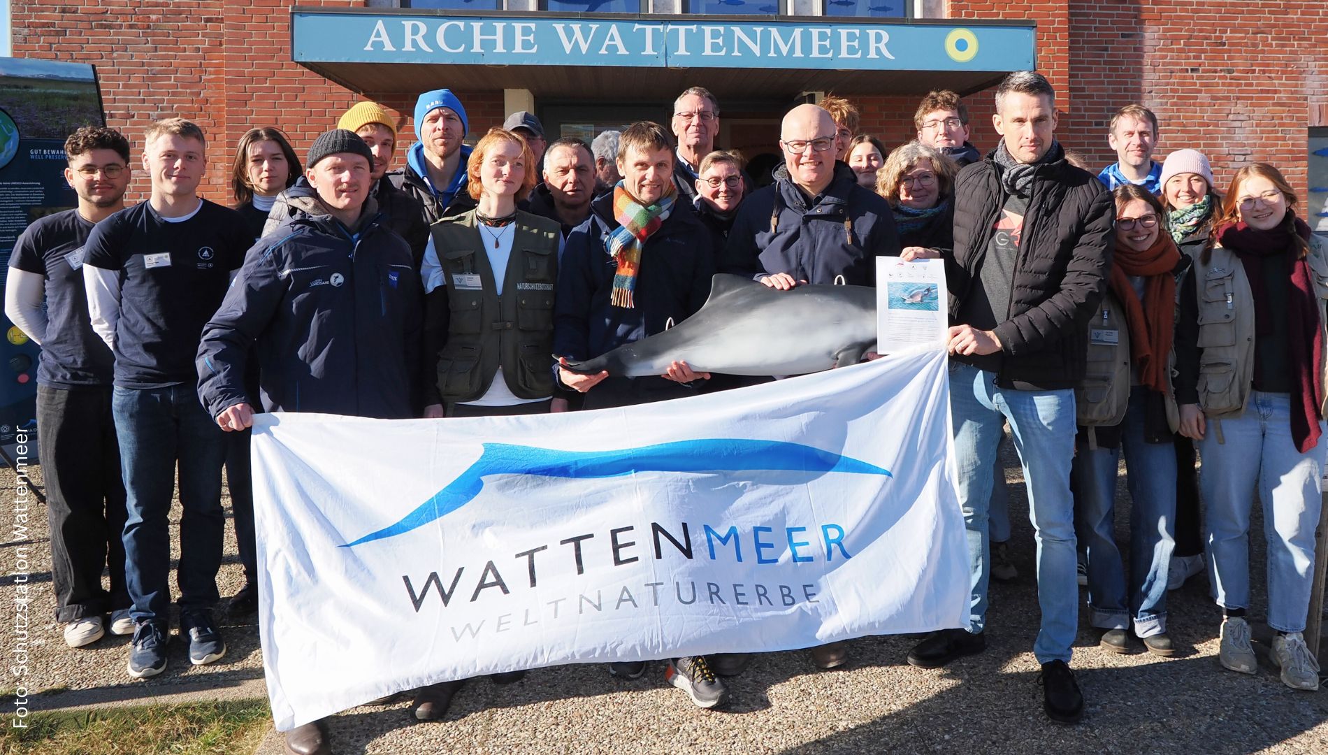 Eine Gruppe von etwa zwanzig Personen steht bei sonnigem Wetter vor dem Gebäude „Arche Wattenmeer“. Sie halten ein großes weißes Banner mit der Aufschrift „Wattenmeer Weltnaturerbe“ und einem stilisierten blauen Wal. Zwei Männer in der Mitte präsentieren ein Modell eines Schweinswals, einer davon hält zusätzlich ein Papier mit einem Foto. 