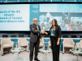 A man and a woman in business attire shake hands and smile while standing on a stage at a formal event. Behind them is a large screen displaying the event title, &ldquo;Launch of the All-Atlantic Network of Coastal Resilience Beacon Sites.