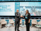 A man and a woman in business attire shake hands and smile while standing on a stage at a formal event. Behind them is a large screen displaying the event title, &ldquo;Launch of the All-Atlantic Network of Coastal Resilience Beacon Sites.