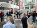 A group of people are gathered in a sunlit indoor atrium for a presentation or discussion. The audience stands in a semicircle around a few tall cocktail tables, attentively listening to a woman speaking near a set of scientific posters. 