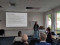 A woman is giving a presentation in a well-lit conference room with large windows. She stands at a podium with a laptop, gesturing as she speaks to an audience seated in front of her.