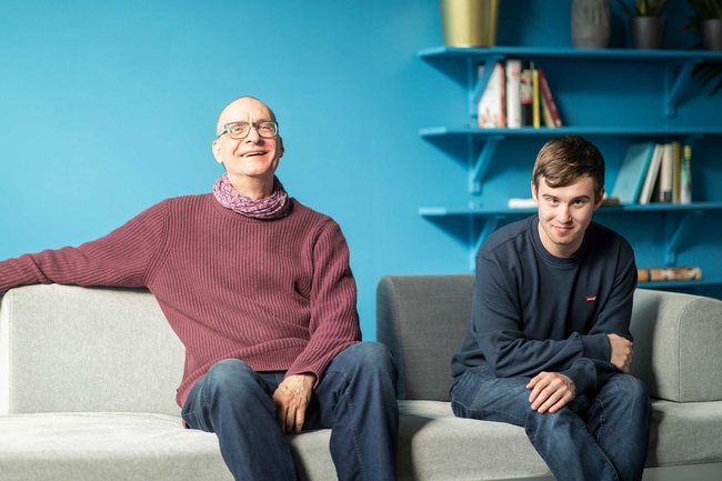 Zwei Personen sitzen auf einem hellgrauen Sofa, links eien Mann mit Brille und einem weinroten Pullover, rechts ein Jugendlicher mit einem blauen Pulli und Jeans. Im Hintergrund sidn eien blaue wan udn eien Bücheregal zu sehen.