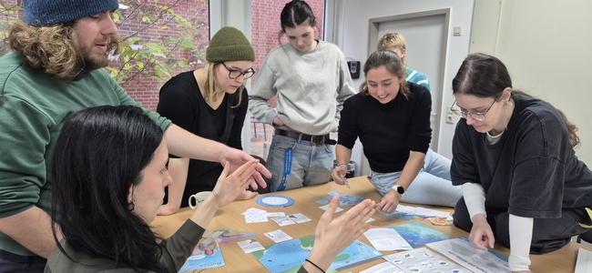 Sieben Erwachsene stehen und sitzen um einen Tisch, der mit blauen Karten, ausgedruckten Blättern und kreisförmigen Diagrammen bedeckt ist. Eine Person gestikuliert mit beiden Händen, während die anderen sich über den Tisch beugen und die Materialien aufmerksam betrachten. Die Gruppe wirkt engagiert in einer gemeinsamen Diskussion oder Arbeitsphase in einem hellen Raum.