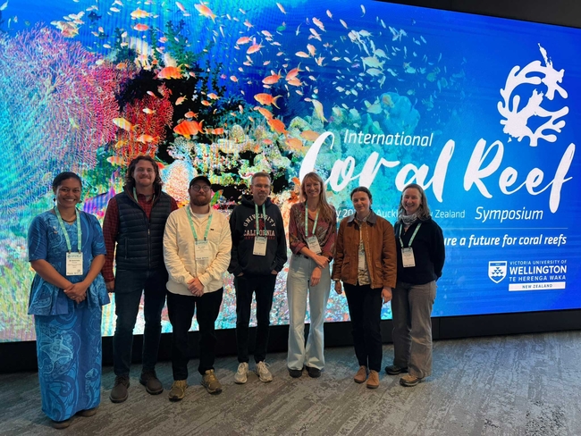 Sieben Personen stehen nebeneinander für ein Gruppenfoto vor einer großen, farbenfrohen Bildschirmwand mit einem Korallenriff und zahlreichen Fischen. Auf dem Bildschirm steht „International Coral Reef Symposium“.
