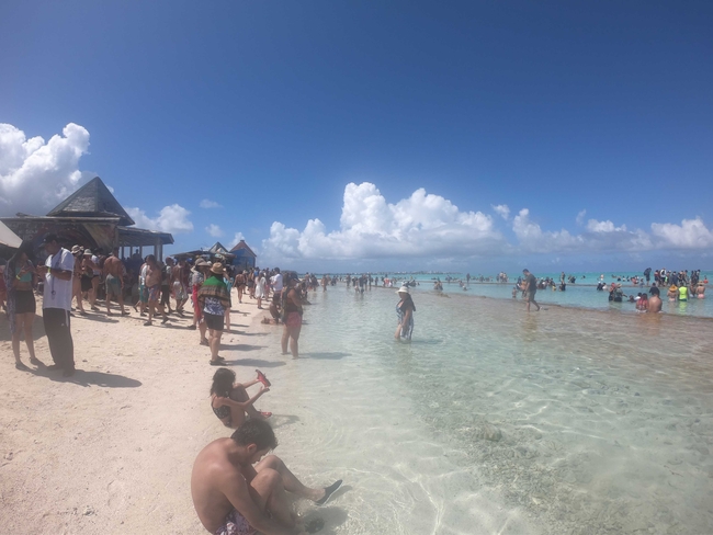 Eine belebte tropische Strandszene mit Dutzenden von Menschen, die einen sonnigen Tag genießen. Viele stehen oder waten im klaren, seichten türkisfarbenen Wasser, während andere am Sandstrand entlanggehen oder sich ausruhen