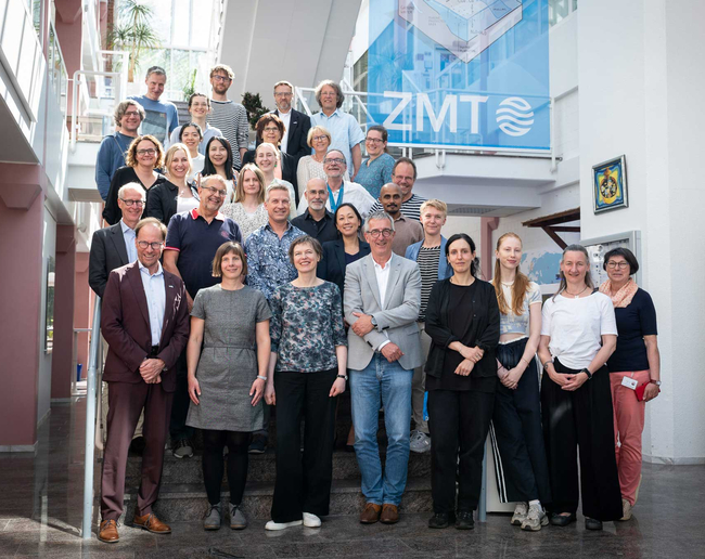 Gruppenfoto von etwa 30 Erwachsenen auf einer breiten Treppe in einem hellen, modernen Atrium mit großen Fenstern und weißen Wänden. Die Personen stehen in mehreren Reihen auf den Stufen und auf einer oberen Galerie, blicken in die Kamera und lächeln. Im Hintergrund hängt ein großes blaues Banner mit der Aufschrift „ZMT“.